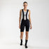A woman wearing black bib shorts with white straps, designed for cycling. The shorts have a high waist and feature a magnetic clip and reflective markings.